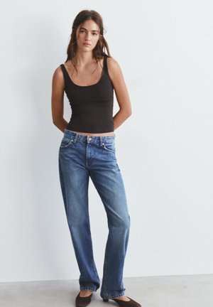 Mladá žena stojaca pri bielej stene, oblečená v čiernom tielku, modrých džínsach s priamym strihom a čiernych balerínach, s rukami za chrbtom.