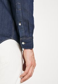 Vänster hand hänger vid sidan, klädd i en mörkblå denimskjorta och vita byxor, mot en enfärgad ljus bakgrund.