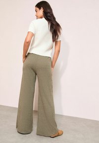 Balts teksturētais īsajām piedurknēm tops, kas pāris ar augstu jostasvietu, ribotām olīvu zaļām platām biksēm. Apavi ir bēši ar siksniņu akcentu.