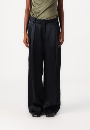 Person trägt hoch taillierte, weit geschnittene schwarze Satin-Hose, kombiniert mit einem in die Hose gesteckten olivgrünen Oberteil, vor weißem Hintergrund stehend.
