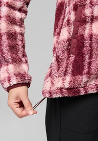 Mano che regola il cordoncino su una giacca in pile a fantasia rosa e bordeaux con tasca laterale con zip, indossata sopra pantaloni neri.