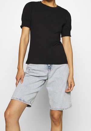 Osoba ubrana w czarną bluzkę z bufkami na rękawach oraz jasnoniebieskie dżinsowe spodenki sięgające do kolan na jednolitym białym tle.