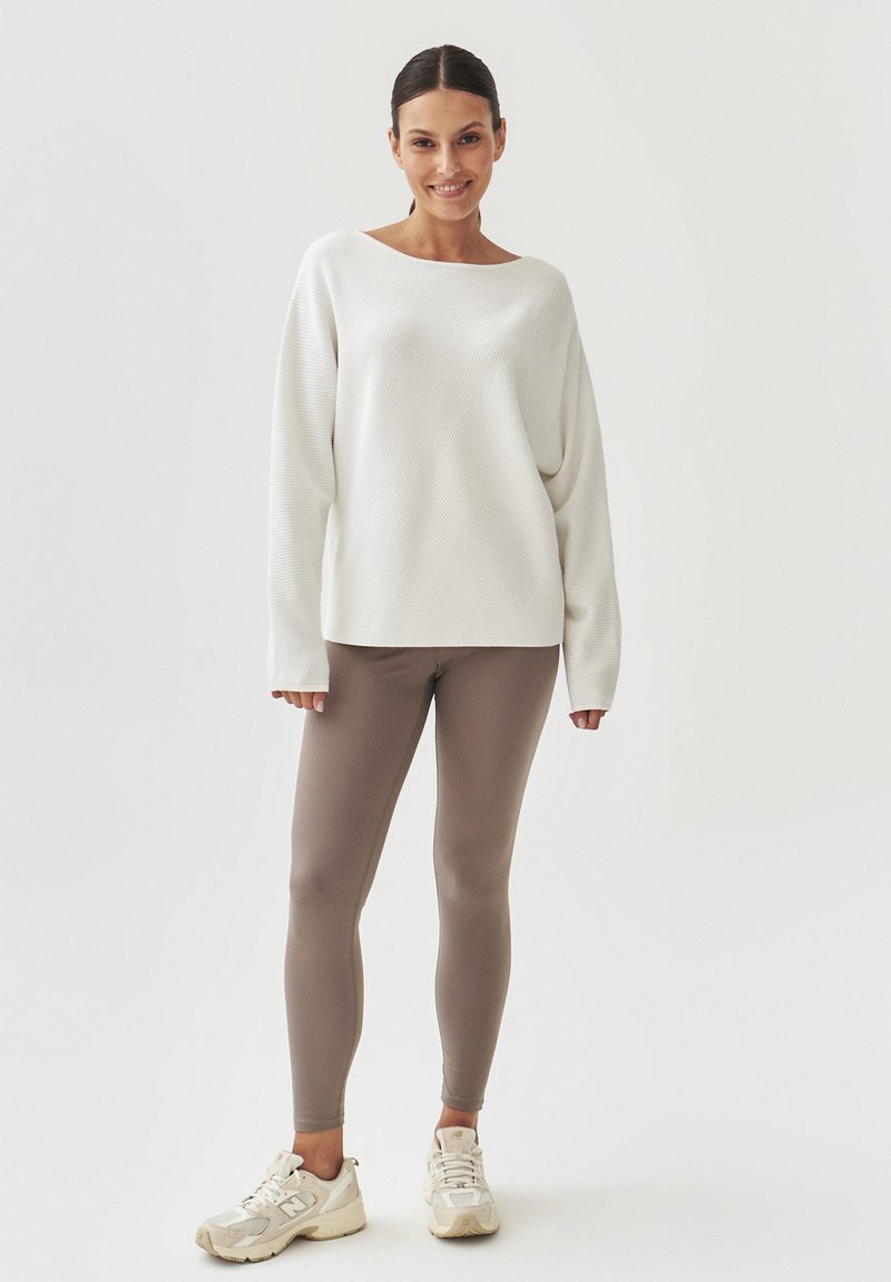 Biały sweter z prążkowanej dzianiny z opadającymi ramionami, w połączeniu z beżowymi legginsami. Modelka nosi kremowe trampki. Jednolity kolor i luźny krój.