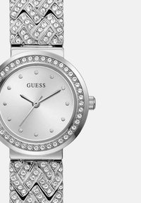 Sølvmetallurvæg med rund skive, udsmykket med krystaller, der har en minimalistisk urskive og et tekstureret chevronmønsterbånd.