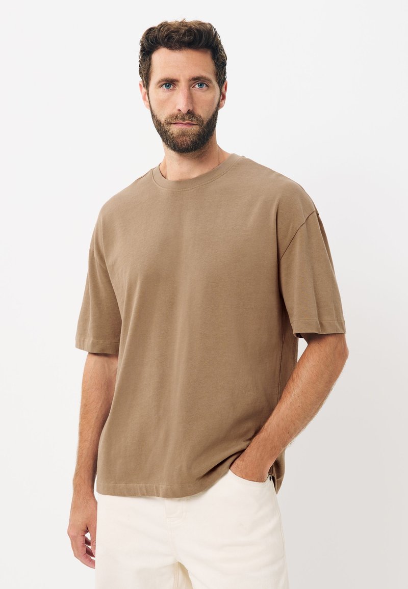 Uomo con capelli corti e ricci e barba, che indossa una camicia marrone a maniche corte e ampia e pantaloni bianchi, in piedi con una mano in tasca.