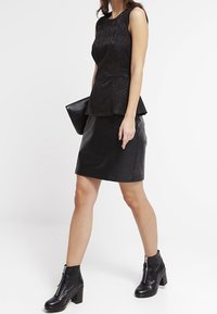 Top peplum nero in pizzo testurizzato abbinato a una gonna in pelle aderente e stivali ankle. Modella che tiene una pochette nera.