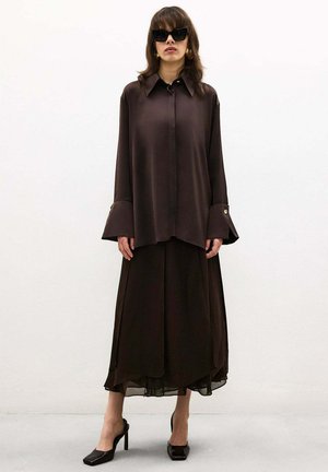 Femme portant une chemise boutonnée marron foncé surdimensionnée, une jupe fluide assortie, des chaussures à talons hauts noires et de grandes lunettes de soleil noires, debout contre un mur blanc.