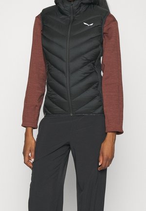 Personne portant un gilet sans manches matelassé noir par-dessus un haut marron à manches longues et un pantalon noir, debout devant un fond uni.