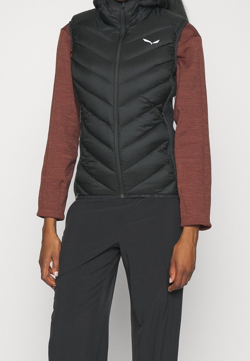 Personne portant un gilet sans manches matelassé noir par-dessus un haut marron à manches longues et un pantalon noir, debout devant un fond uni.