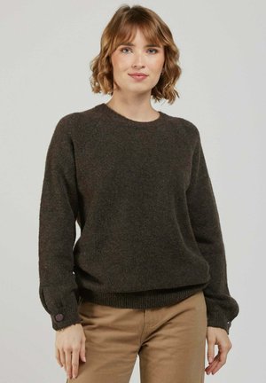 Femme aux cheveux courts et ondulés portant un pull brun foncé avec des poignets boutonnés et un pantalon beige, debout devant un fond clair uni.
