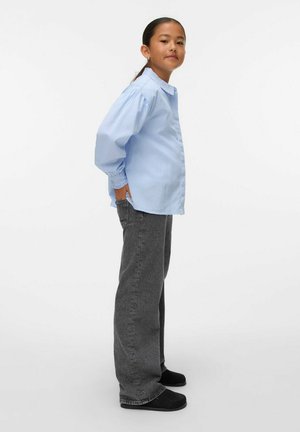 Camisa de botones de manga larga a rayas azules y blancas, jeans anchos grises, zapatos negros sin cordones, pose de pie, manos en los bolsillos.
