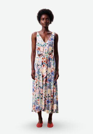 Femme aux cheveux courts et bouclés portant une robe sans manches à motif coloré avec une ceinture à la taille et des chaussures rouges en tissu tressé, debout devant un fond blanc.