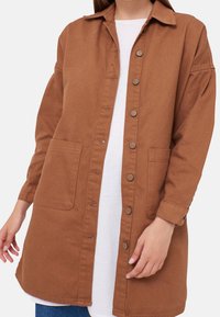 Žena v dlouhém hnědém kabátu na knoflíky s dvěma předními kapsami, oblečená přes bílé tričko a modré džíny, zachycena od krku po polovinu stehna.