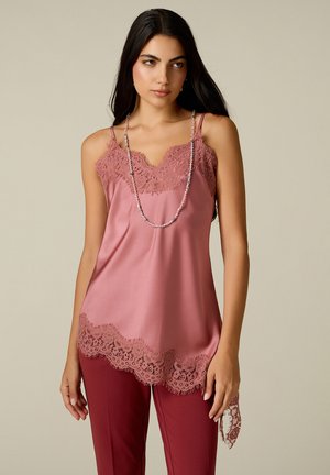 Mujer con cabello largo y oscuro que lleva una camisola rosa con borde de encaje y pantalones rojo oscuro, de pie frente a un fondo neutro.