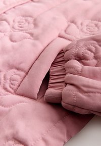 Lyserødt quiltet stof med blomstret syning og en lomme, der viser samlet elastisk detalje på kanten af tekstilet.