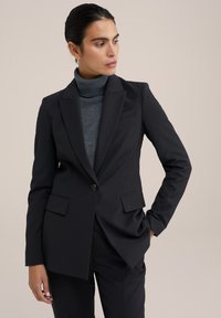Schwarzer taillierter Blazer mit Revers, Ein-Knopf-Verschluss und zwei Fronttaschen. Getragen über einem schmalen grauen Rollkragenpullover.