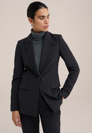 Blazer nero su misura con rever a punta, chiusura a un bottone e due tasche frontali. Indossato sopra un dolcevita grigio aderente.