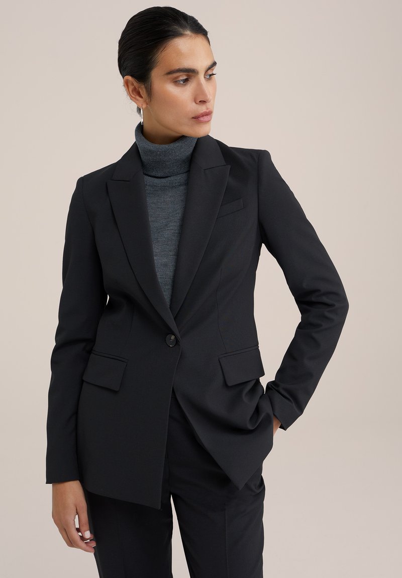 Schwarzer taillierter Blazer mit Revers, Ein-Knopf-Verschluss und zwei Fronttaschen. Getragen über einem schmalen grauen Rollkragenpullover.