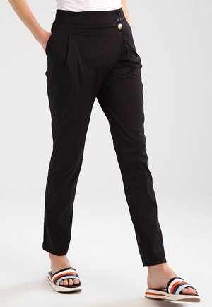 Kobieta ubrana w czarne, zwężane spodnie z wysokim stanem z trzema guzikami na talii, połączone z czarnymi klapkami w paski w kolorach czarnym, białym i pomarańczowym.