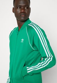 Zaļš zip-up jaciņa ar baltām svītrām gar pleciem un piedurknēm; ar uzceltu apkakli un adidas logo uz krūtīm.