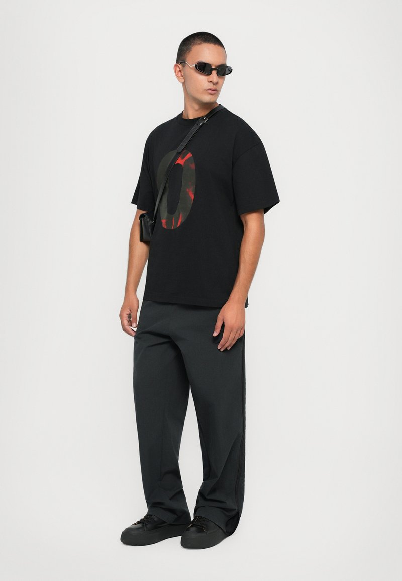 Black oversized t-shirt with circular olive and red graphic, paired with black wide trousers and black sneakers. Accessories include small bag and sunglasses.