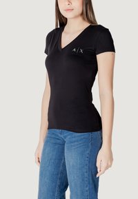 T-shirt in cotone nero con scollo a V, maniche corte e design aderente. Presenta un sottile logo A/X sul petto. Indossato con jeans blu.