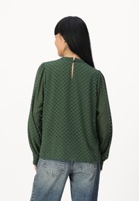 Blouse vert foncé à motifs avec une finition texturée, manches longues, et une ouverture en keyhole au dos fermée par un bouton. Portée avec un jean bleu clair.