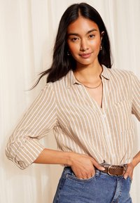 Šviesiai ruda ir balta dryžuota marškinėlių palaidinė su sagomis, apykakle, užlenktomis rankovėmis ir krūtinės kišene, derinama su aukšto liemens mėlynais džinsais ir ruduoju diržu.