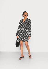 Vestido preto e branco com padrão, mangas longas, decote em V e design de tabuleiro de xadrez. Acompanhado por uma mala preta e sapatos de salto aberto.