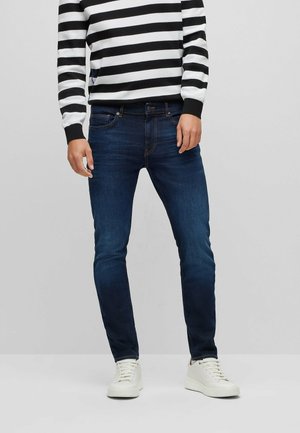Hombre vestido con jeans ajustados de color azul oscuro, zapatillas blancas y una camisa de manga larga a rayas en blanco y negro, de pie contra un fondo liso.
