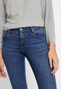 Mörkblå jeans med en smal passform, utrustade med två silverfärgade knapplås, sydda fickor och subtila blekningsdetaljer på tyget.