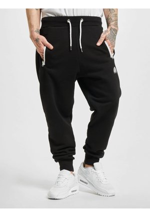 Juodi joggeriai, pagaminti iš minkštos medžiagos, su elastinėmis rankovėmis, reguliuojamu raišteliu ir užtrauktukais šoninėse kišenėse. Matomas baltas logotipo detalė.