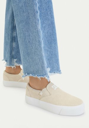 Osoba nosząca jasnoniebieskie jeansy z postrzępionymi nogawkami oraz beżowe, wsuwane sneakersy na platformie na białym tle.