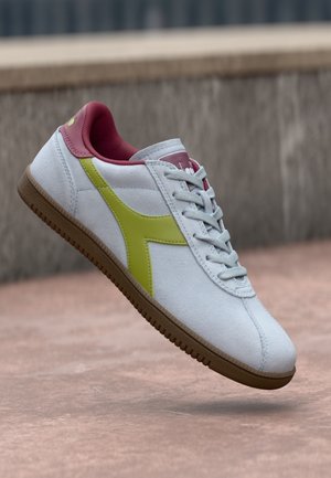 White sneaker with a yellow side stripe and pink accents. Features textured material, a lace-up design, and a brown rubber sole.