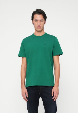 Man wearing green short-sleeve t-shirt with small black logo on chest, paired with dark blue jeans, standing against white background.