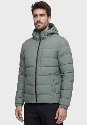 Mann trägt eine grüne wattierte Jacke mit Kapuze, steht vor einem einfachen hellen Hintergrund und blickt leicht zur Seite.