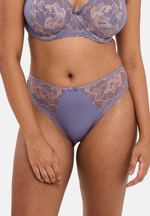 Mujer con sujetador de encaje púrpura y braguitas de encaje a juego de corte alto, de pie con los brazos relajados a los lados.