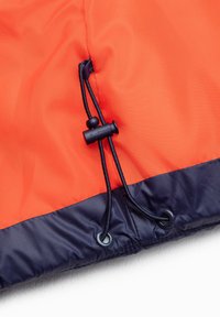 Details van de oranje en marineblauwe waterdichte jas met trekkoord en zwarte koord, gekenmerkt door een gladde textuur en gestikte randen.