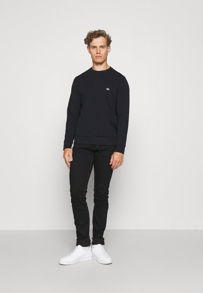 Felpa nera a collo rotondo con polsini a costine, abbinata a jeans neri slim e sneakers bianche. Il modello si trova su uno sfondo neutro.