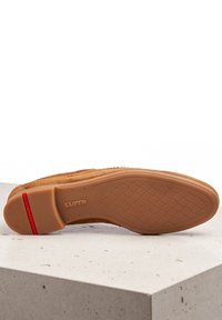 Chaussure en daim tan avec une semelle en caoutchouc texturé, dotée d'une bande rouge et de coutures détaillées le long du bord. Le design de la semelle met en avant un motif de traction.