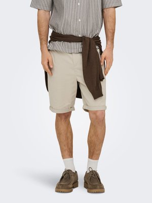 Mann trägt beige hochgekrempelte Shorts, gemustertes kurzärmeliges Hemd, braunen Pullover um die Taille gebunden, weiße Socken und braune Wildlederschuhe.