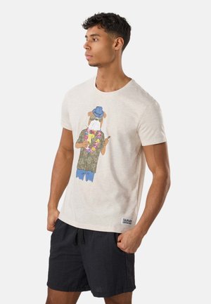 Young man wearing beige t-shirt with dog in tropical outfit graphic and black shorts, standing with hands in pockets, looking left.