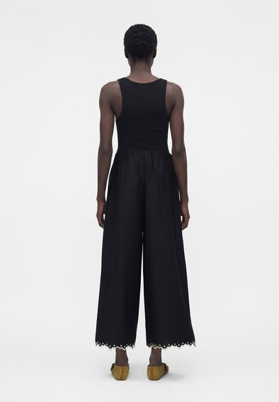 Άτομο που στέκεται με την πλάτη γυρισμένη, φοράει μαύρο αμάνικο τοπ, φαρδιά μαύρα παντελόνια με κυματιστά τελειώματα και κίτρινα πλεκτά παπούτσια χωρίς κορδόνια.