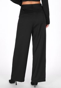 Pantaloni negri cu croi larg dintr-un material neted, având două buzunare pe spate și un tiv structurat, asortați cu pantofi cu toc ascuțit negri.