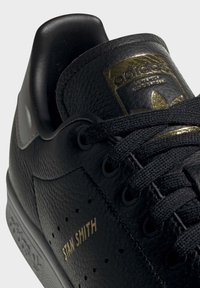 Schwarze Ledersneaker mit strukturierten Oberflächen, goldenen Akzenten und einem klassischen Stan Smith-Label. Schwarze Schnürsenkel und ein gepolsterter Knöchelkragen.