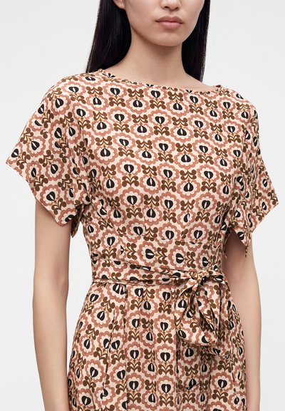 Femme portant une robe à manches courtes avec une ceinture à nouer à la taille et un motif floral répétitif dans des tons de rose, marron et noir sur tissu beige.