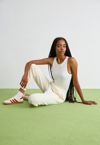 Witte geribbelde tanktop met hoge hals, crèmekleurige joggers en beige sneakers met rode strepen, zittend op een groen tapijt met een neutrale achtergrond.