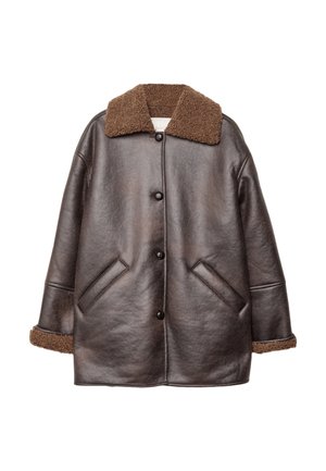 Persoon die een donkerbruine leren jas draagt met een getextureerde bruine shearling kraag en manchetten, met gouden ringen en een ketting met een bloemenhanger.