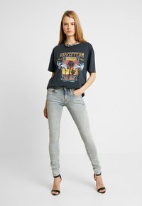 T-shirt gráfica preta com um design do Led Zeppelin, combinado com jeans skinny cinza claro e sandálias de salto pretas. Acento de corrente no colar.