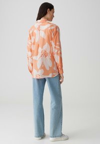 Camicia a maniche lunghe leggera in corallo con ampia stampa floreale bianca. Presenta un colletto classico e polsini con bottone, abbinata a jeans di denim azzurro chiaro.